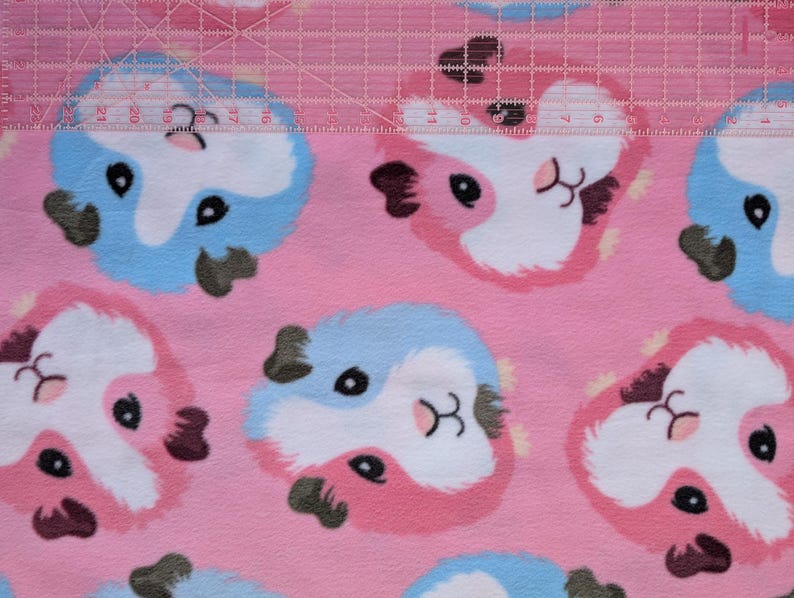 May include: Pink fabric with a repeating pattern of cartoon guinea pig faces in pink, white, blue, and black. A clear ruler with white markings is laid across the top of the fabric.