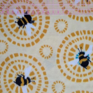 May include: A light yellow fabric with a repeating pattern of stylized bees and concentric circles in shades of yellow and orange. The bees are black and yellow with white wings. The circles have a dotted design.