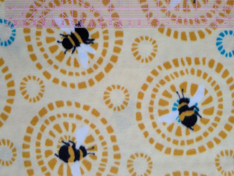 May include: A light yellow fabric with a repeating pattern of stylized bees and concentric circles in shades of yellow and orange. The bees are black and yellow with white wings. The circles have a dotted design.