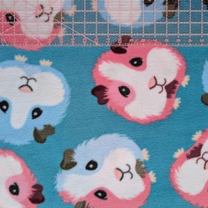 May include: Close-up of a blue fabric with a repeating pattern of cartoon guinea pigs in pink, white, and blue. The guinea pigs have black eyes and brown ears. A clear ruler with inch markings is across the top.