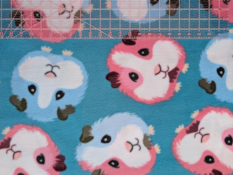May include: Close-up of a blue fabric with a repeating pattern of cartoon guinea pigs in pink, white, and blue. The guinea pigs have black eyes and brown ears. A clear ruler with inch markings is across the top.