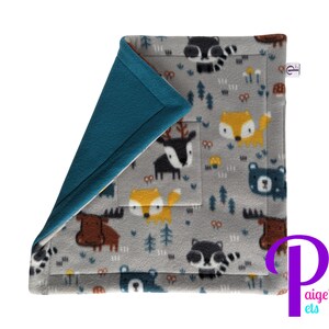 May include: A gray fleece blanket with a forest animal pattern, including foxes, bears, raccoons, and moose. The blanket has a teal blue border and a tag that says "paige's els".