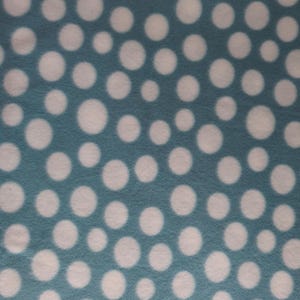 May include: Close-up of a teal fabric with a pattern of white polka dots of varying sizes. The fabric has a soft, fuzzy texture. The polka dots are evenly spaced across the surface.