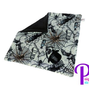 May include: A square pet blanket with a white background featuring a pattern of black spiders, spiderwebs, bats, skulls wearing top hats, and tombstones with RIP text. The reverse side of the blanket is solid black fabric, and a small white tag is visible.