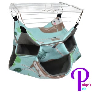 May include: A multi-level pet hammock in a light blue fabric with a sloth pattern. The hammock has three compartments and is designed to hang from a cage. The interior is gray, and the hammock is suspended by black straps.