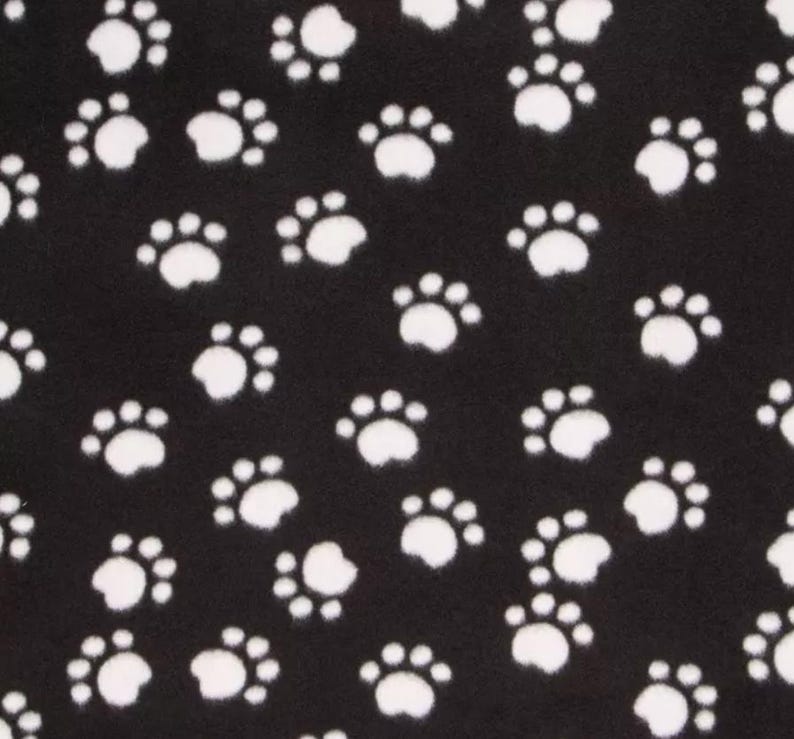 May include: Black fabric with a repeating pattern of white paw prints. The paw prints are scattered across the surface, creating a playful and whimsical design. The fabric appears to be soft and textured.