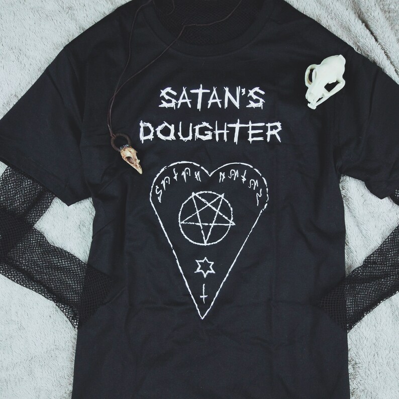 May include: Black t-shirt with the text "Satan's Daughter" in white lettering. A pentagram inside a heart shape is printed below the text.