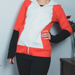 Puede incluir: Una sudadera con capucha roja, blanca y negra con cremallera. La sudadera tiene un panel blanco en la parte delantera y paneles rojos en los laterales.