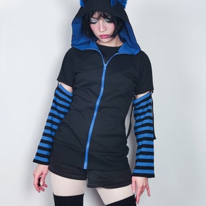 Puede incluir: Una sudadera con capucha negra con detalles azules y cierre de cremallera. La capucha tiene un par de orejas de gato azules en la parte superior. Las mangas son de rayas azules y negras.