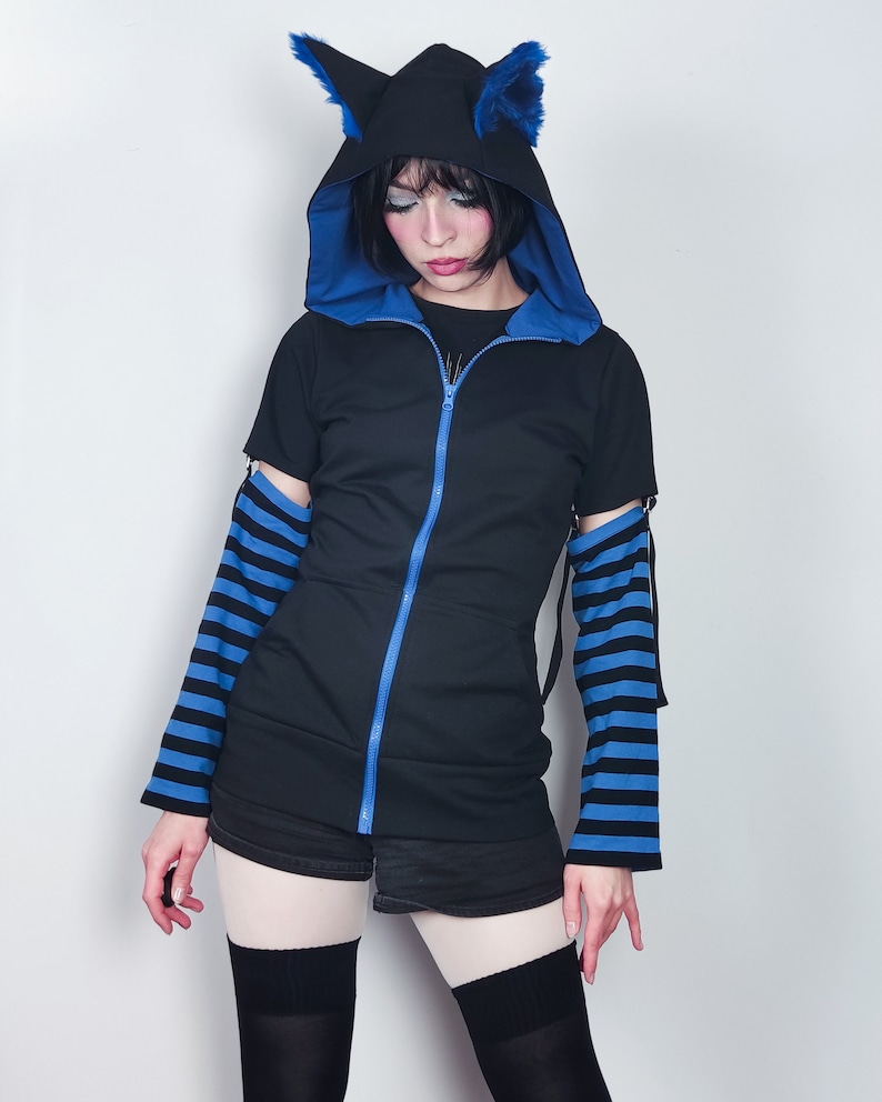 Puede incluir: Una sudadera con capucha negra con detalles azules y cierre de cremallera. La capucha tiene un par de orejas de gato azules en la parte superior. Las mangas son de rayas azules y negras.
