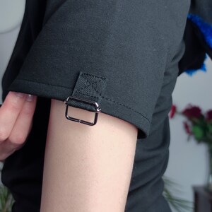 Puede incluir: Camiseta negra de manga corta con un detalle de hebilla plateada en la manga.