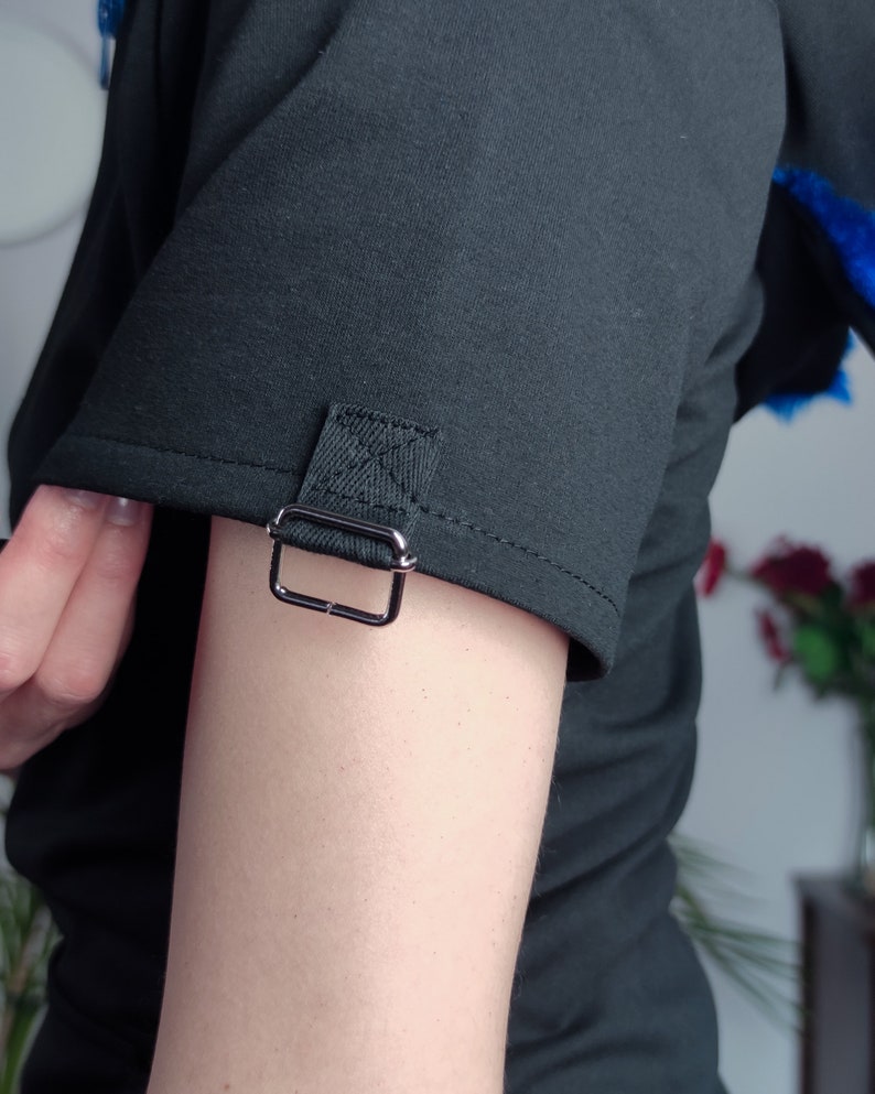 Puede incluir: Camiseta negra de manga corta con un detalle de hebilla plateada en la manga.