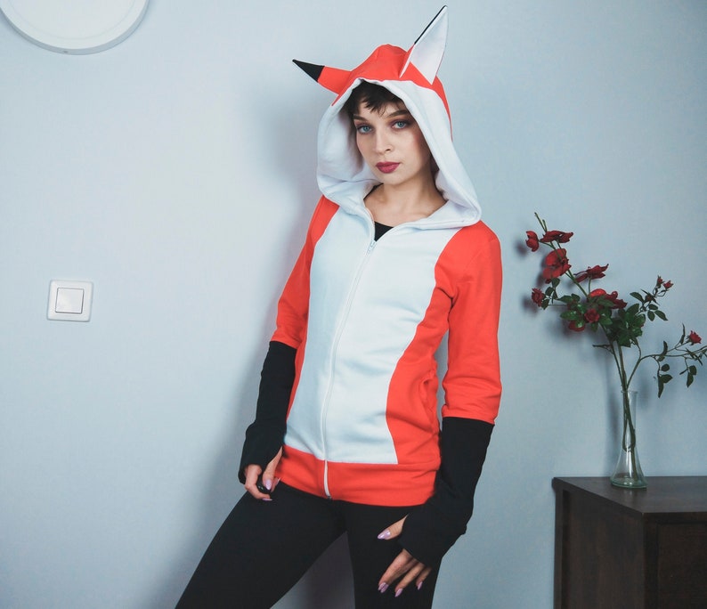 Puede incluir: Una sudadera con capucha blanca y naranja con un dise&ntilde;o de zorro. La capucha tiene orejas grandes y un forro blanco. La sudadera tiene una cremallera y una cintura negra.