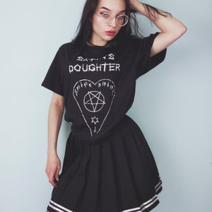 May include: A black t-shirt with the text "SATAN'S DAUGHTER" and a pentagram inside a heart. The person is wearing a black pleated skirt with white stripes and black fishnet stockings.