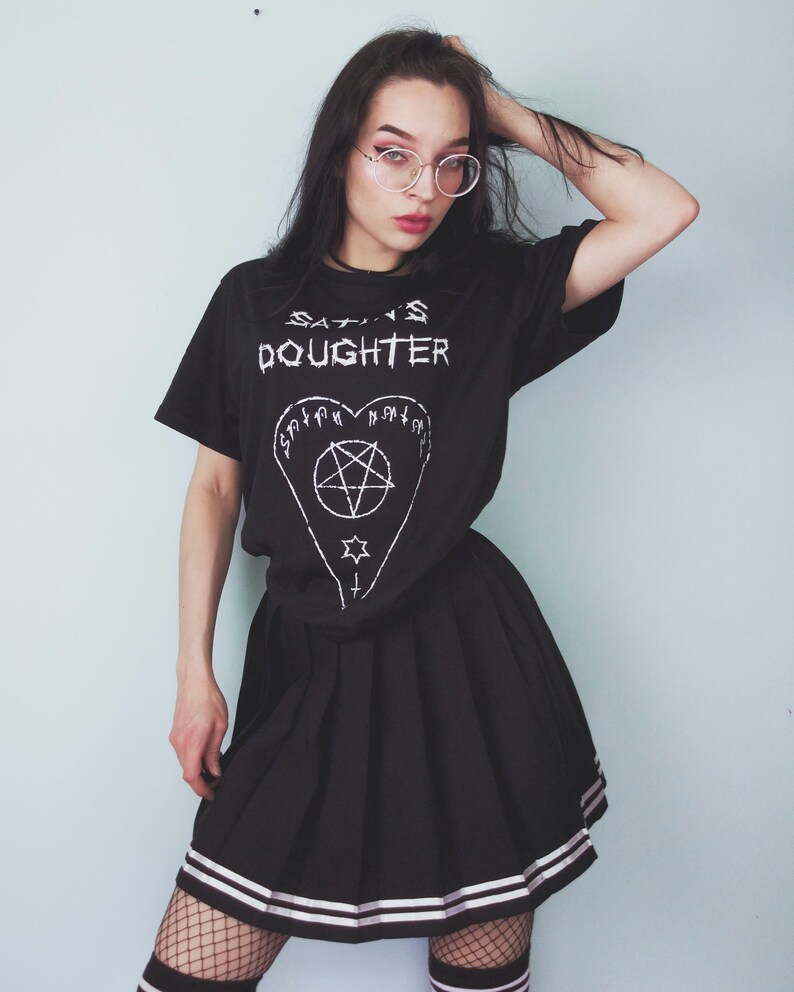 May include: A black t-shirt with the text "SATAN'S DAUGHTER" and a pentagram inside a heart. The person is wearing a black pleated skirt with white stripes and black fishnet stockings.