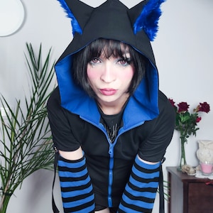 Puede incluir: Una sudadera con capucha negra con orejas de gato de piel azul, ribetes azules y mangas a rayas azules y negras. La cremallera est&aacute; parcialmente abierta.
