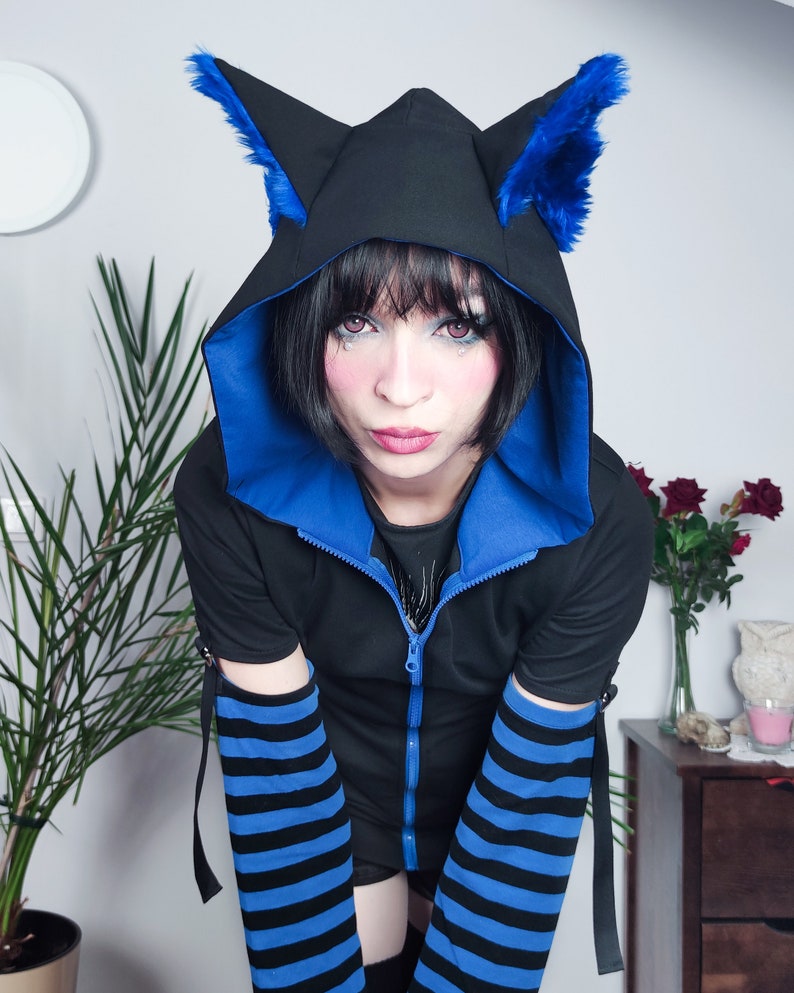 Puede incluir: Una sudadera con capucha negra con orejas de gato de piel azul, ribetes azules y mangas a rayas azules y negras. La cremallera est&aacute; parcialmente abierta.