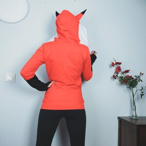 Puede incluir: Una sudadera con capucha roja con un dise&ntilde;o de cabeza de zorro blanco y negro en la capucha. La sudadera la lleva puesta una persona con leggings negros.