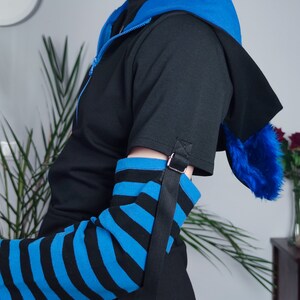 Puede incluir: Una manga a rayas negras y azules con una correa y hebilla negras. La manga se lleva sobre una camisa negra con una chaqueta con capucha azul.