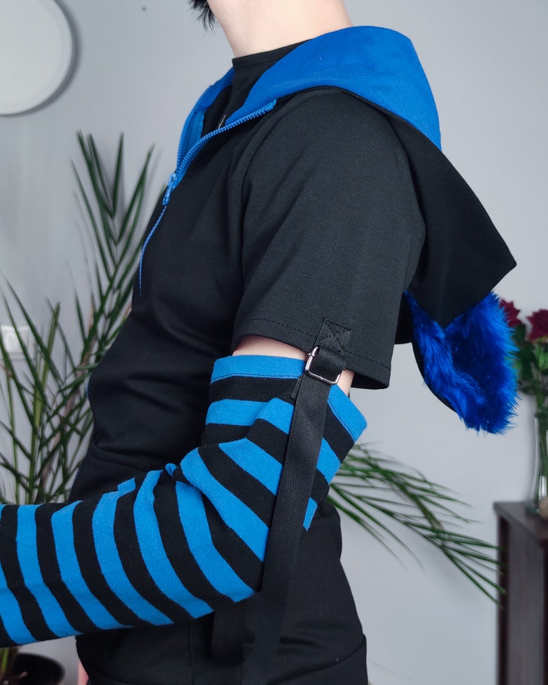 Puede incluir: Una manga a rayas negras y azules con una correa y hebilla negras. La manga se lleva sobre una camisa negra con una chaqueta con capucha azul.