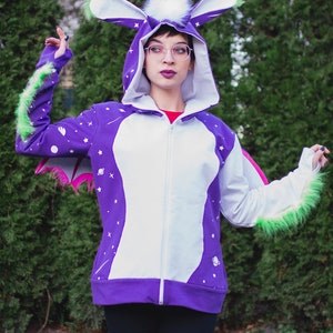 May include: A purple and white hooded sweatshirt with a zipper closure and a white fur trim around the hood. The sweatshirt has a space-themed design with stars and planets. The hood has large, white, furry ears with green tips.