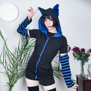 Puede incluir: Una sudadera con capucha negra con orejas de gato de piel azul y mangas a rayas azules y negras. La sudadera tiene una cremallera y se combina con una falda corta y calcetines hasta la rodilla.