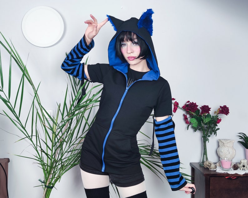 Puede incluir: Una sudadera con capucha negra con orejas de gato de piel azul y mangas a rayas azules y negras. La sudadera tiene una cremallera y se combina con una falda corta y calcetines hasta la rodilla.