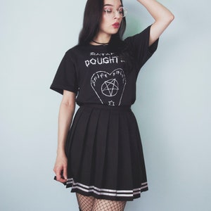 May include: A person wearing a black t-shirt with the text "SATAN  OUGHT TO" and a pentagram design. The person is also wearing a black pleated skirt with white stripes, black fishnet stockings, and black and white striped socks.