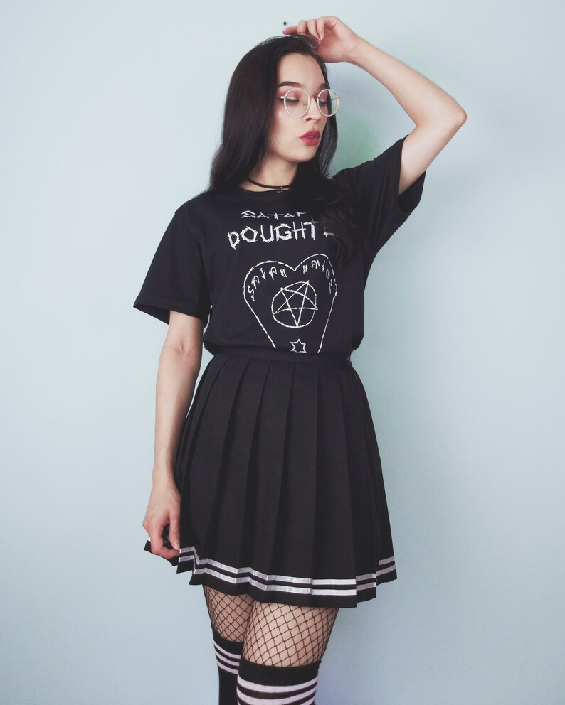 May include: A person wearing a black t-shirt with the text "SATAN  OUGHT TO" and a pentagram design. The person is also wearing a black pleated skirt with white stripes, black fishnet stockings, and black and white striped socks.