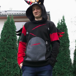 May include: A person wearing a black hoodie with red and grey accents. The hoodie has a large grey animal head design on the front and a pair of red horns on the hood. The person is standing in front of a green bush.