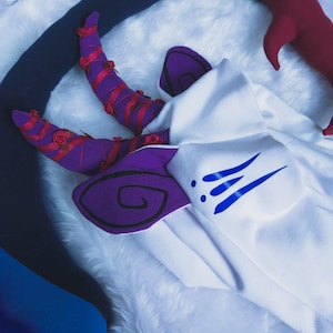 May include: A white plush costume with purple and blue details. The costume features a pair of purple horns with red roses and a blue tail.
