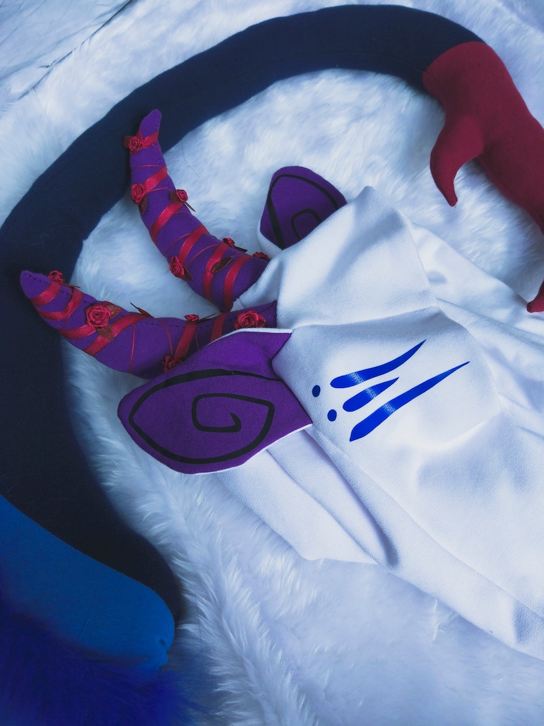 May include: A white plush costume with purple and blue details. The costume features a pair of purple horns with red roses and a blue tail.