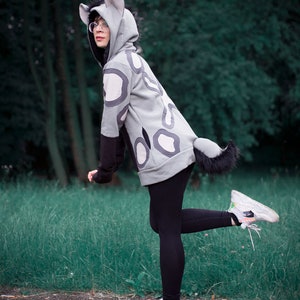 May include: A person wearing a gray hooded sweatshirt with a black and white spotted pattern. The sweatshirt has a furry tail and ears. The person is wearing black leggings and white sneakers.