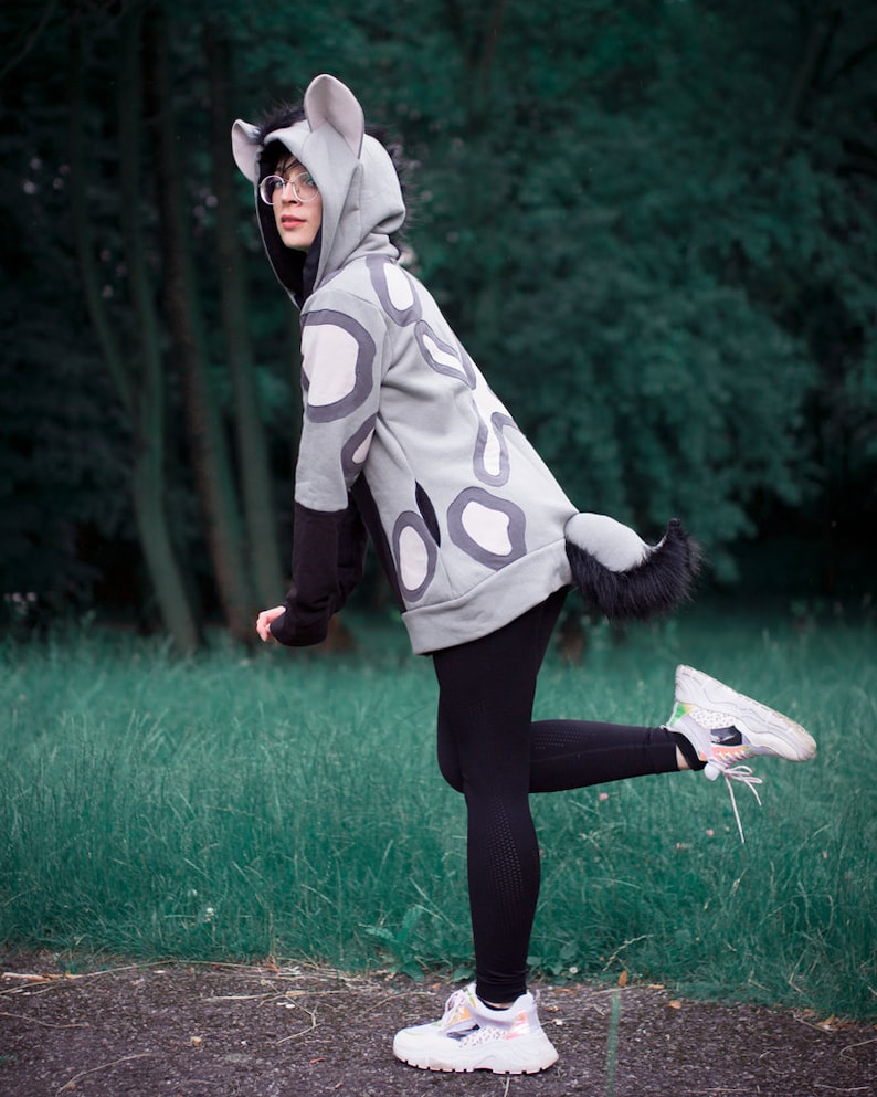 May include: A person wearing a gray hooded sweatshirt with a black and white spotted pattern. The sweatshirt has a furry tail and ears. The person is wearing black leggings and white sneakers.