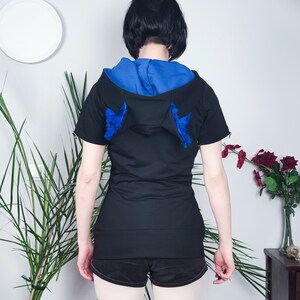 Puede incluir: Sudadera negra de manga corta con capucha azul y orejas de piel azul en la parte posterior.