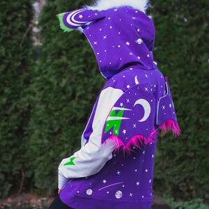 May include: Purple hoodie with a white and green space-themed design. The hoodie has a large hood with white fur trim and a pair of pink wings with green and white accents.