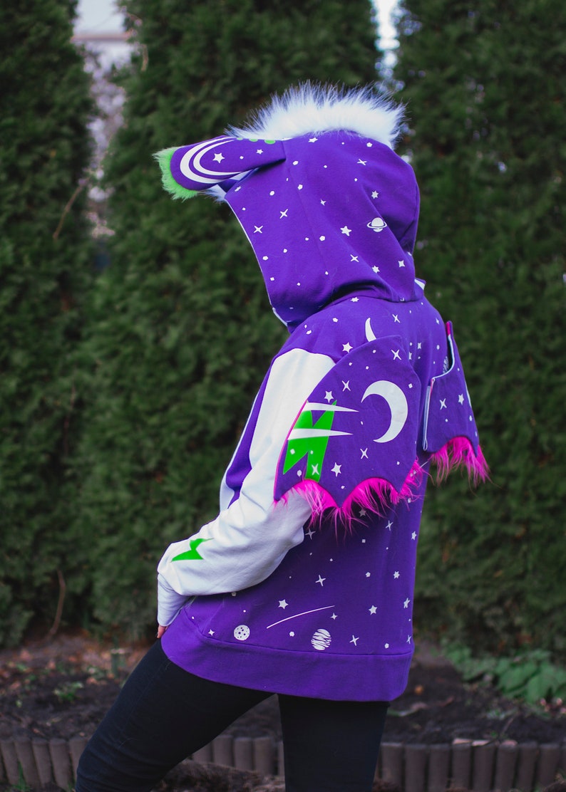 May include: Purple hoodie with a white and green space-themed design. The hoodie has a large hood with white fur trim and a pair of pink wings with green and white accents.