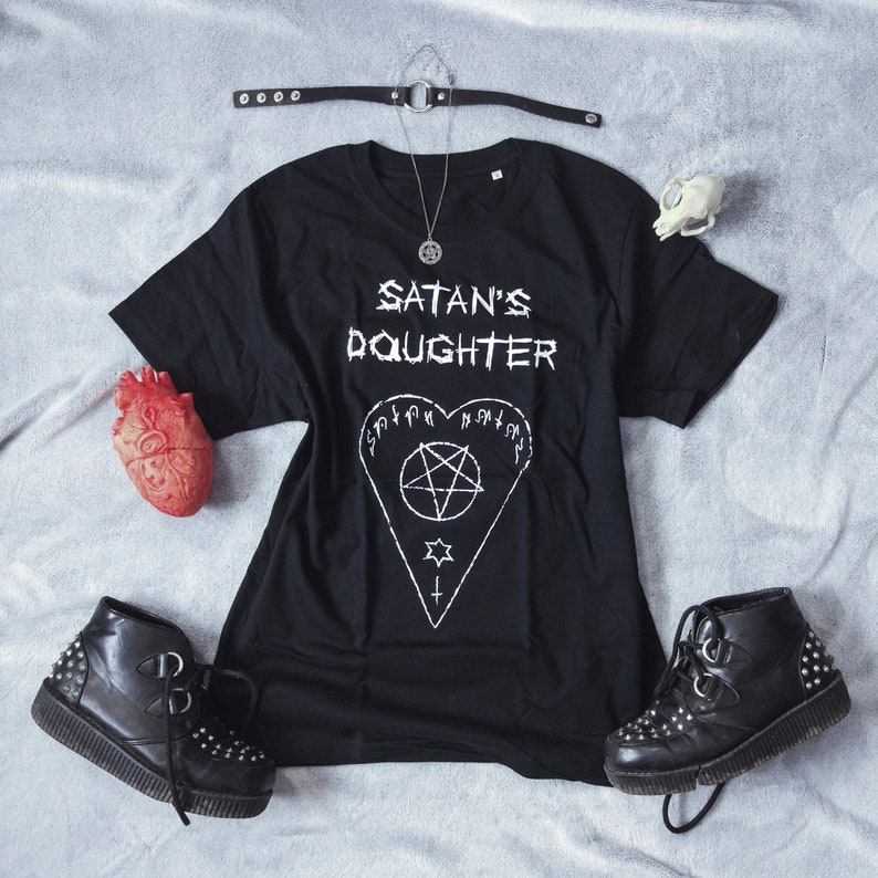 May include: Black t-shirt with the text "SATAN'S DAUGHTER" and a pentagram inside a heart. The shirt is laid out on a gray surface with a silver necklace, a small white skull, and a red heart.