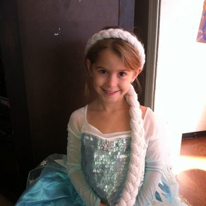 May include: A young girl dressed as Elsa from the movie Frozen. She is wearing a light blue dress with a sparkly bodice and a white braid. She is also wearing a white headband.