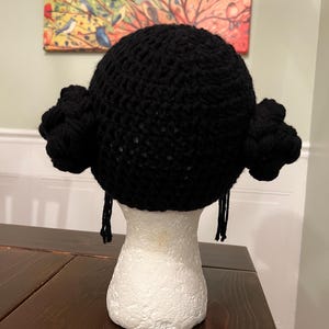 May include: A black crocheted hat with side buns and dangling strands, displayed on a white mannequin head. The hat is made of thick yarn and has a rounded shape. The buns are positioned on either side of the head, and the strands hang down.