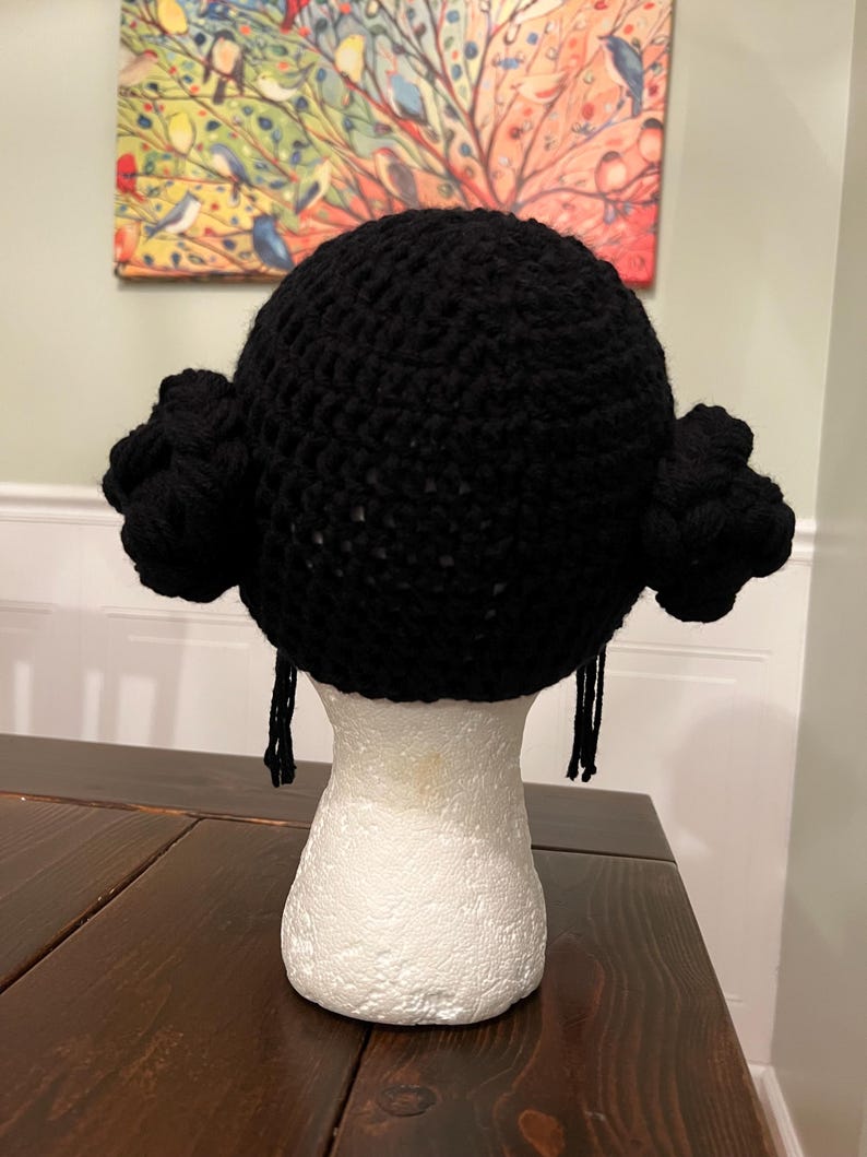 May include: A black crocheted hat with side buns and dangling strands, displayed on a white mannequin head. The hat is made of thick yarn and has a rounded shape. The buns are positioned on either side of the head, and the strands hang down.