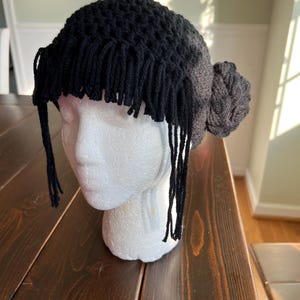 May include: A black crochet hat with fringe and a bun-shaped detail. The hat is displayed on a white mannequin head. The fringe hangs down around the forehead. The bun is on the right side of the head.