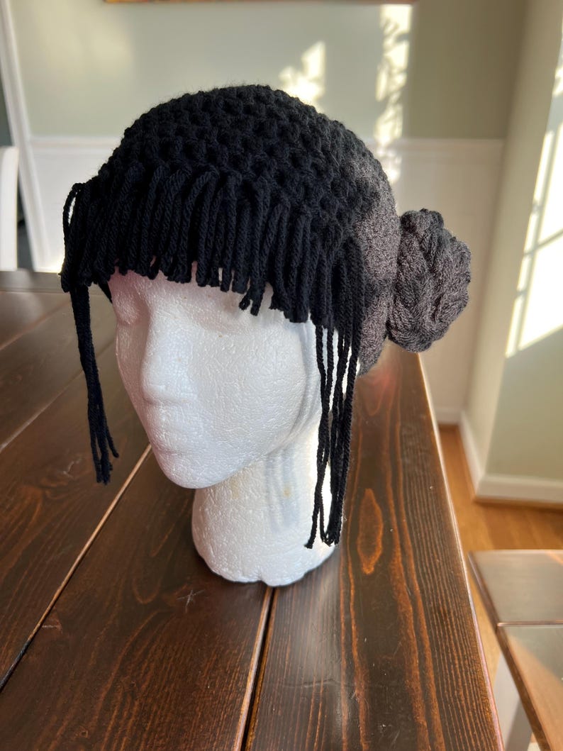 May include: A black crochet hat with fringe and a bun-shaped detail. The hat is displayed on a white mannequin head. The fringe hangs down around the forehead. The bun is on the right side of the head.