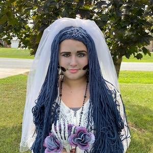 May include: A woman dressed as a bride with blue yarn hair, a white veil, and a white lace top. She is holding a bouquet of purple roses and a skeleton hand.