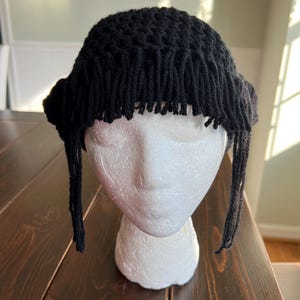 May include: A black crochet hat with fringe detail. The hat is displayed on a white foam head. The hat has a round crown and a fringe that hangs down around the forehead and sides. The hat is made of a thick yarn.