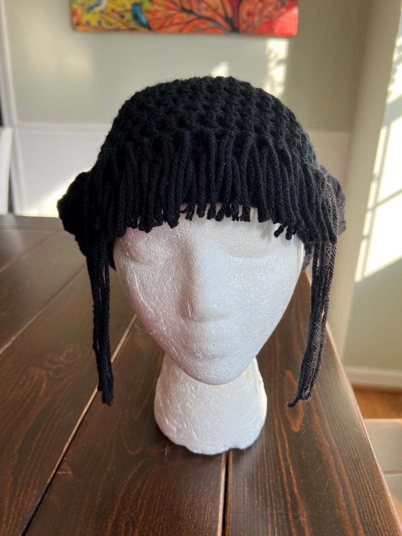 May include: A black crochet hat with fringe detail. The hat is displayed on a white foam head. The hat has a round crown and a fringe that hangs down around the forehead and sides. The hat is made of a thick yarn.