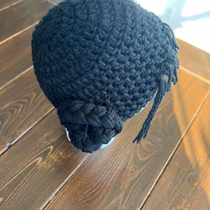 May include: A black crocheted hat with a textured stitch pattern. The hat features a braided accent on the side and fringe detailing. The hat is sitting on a wooden surface.