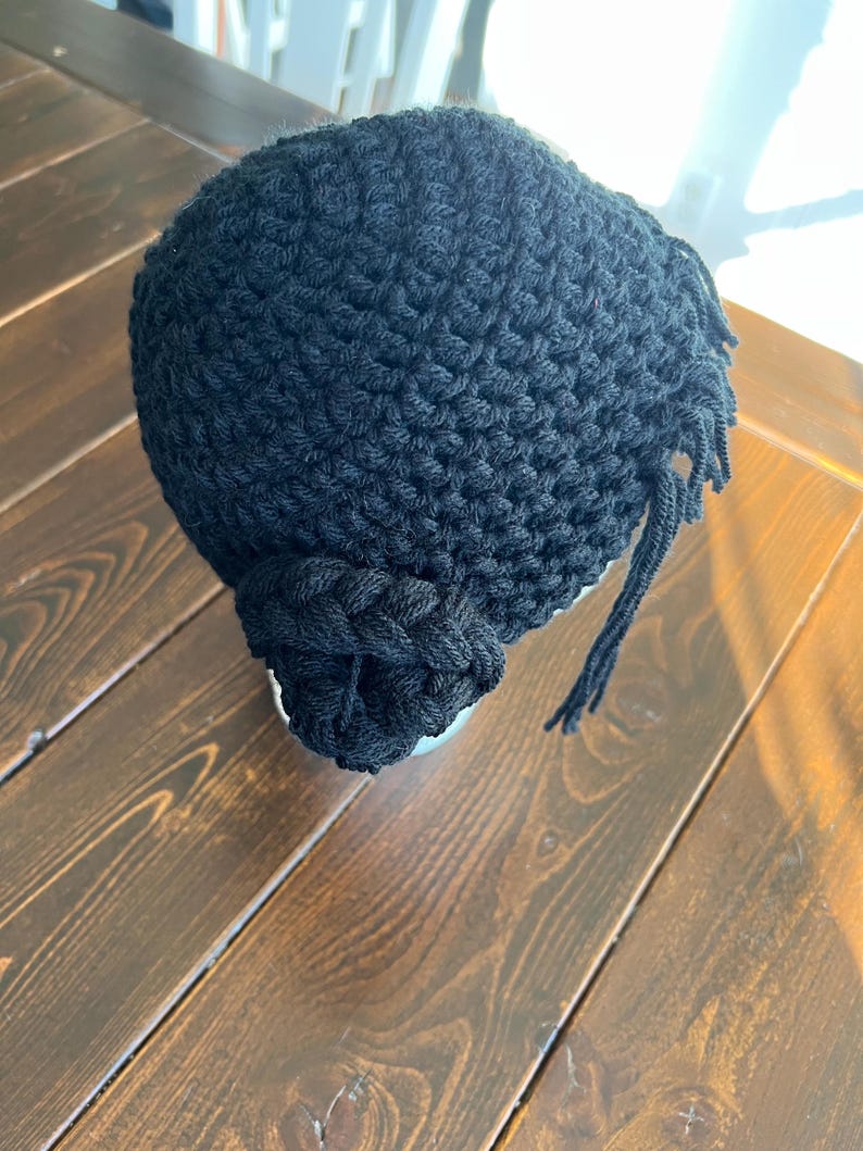 May include: A black crocheted hat with a textured stitch pattern. The hat features a braided accent on the side and fringe detailing. The hat is sitting on a wooden surface.