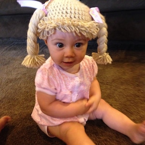May include: A baby wearing a crocheted hat with yarn braids and pink bows. The hat is beige and the braids are blonde.