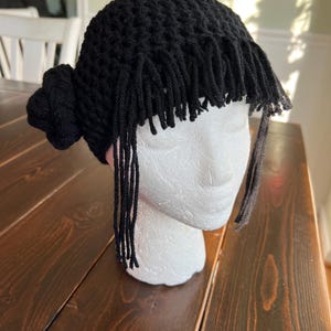 May include: A black crochet hat with fringe and a bun detail. The hat is displayed on a white mannequin head, showcasing the handmade design. The fringe hangs around the forehead and sides.